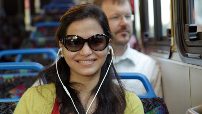 smiling woman listening to music on the bus, man behind her looking out the window